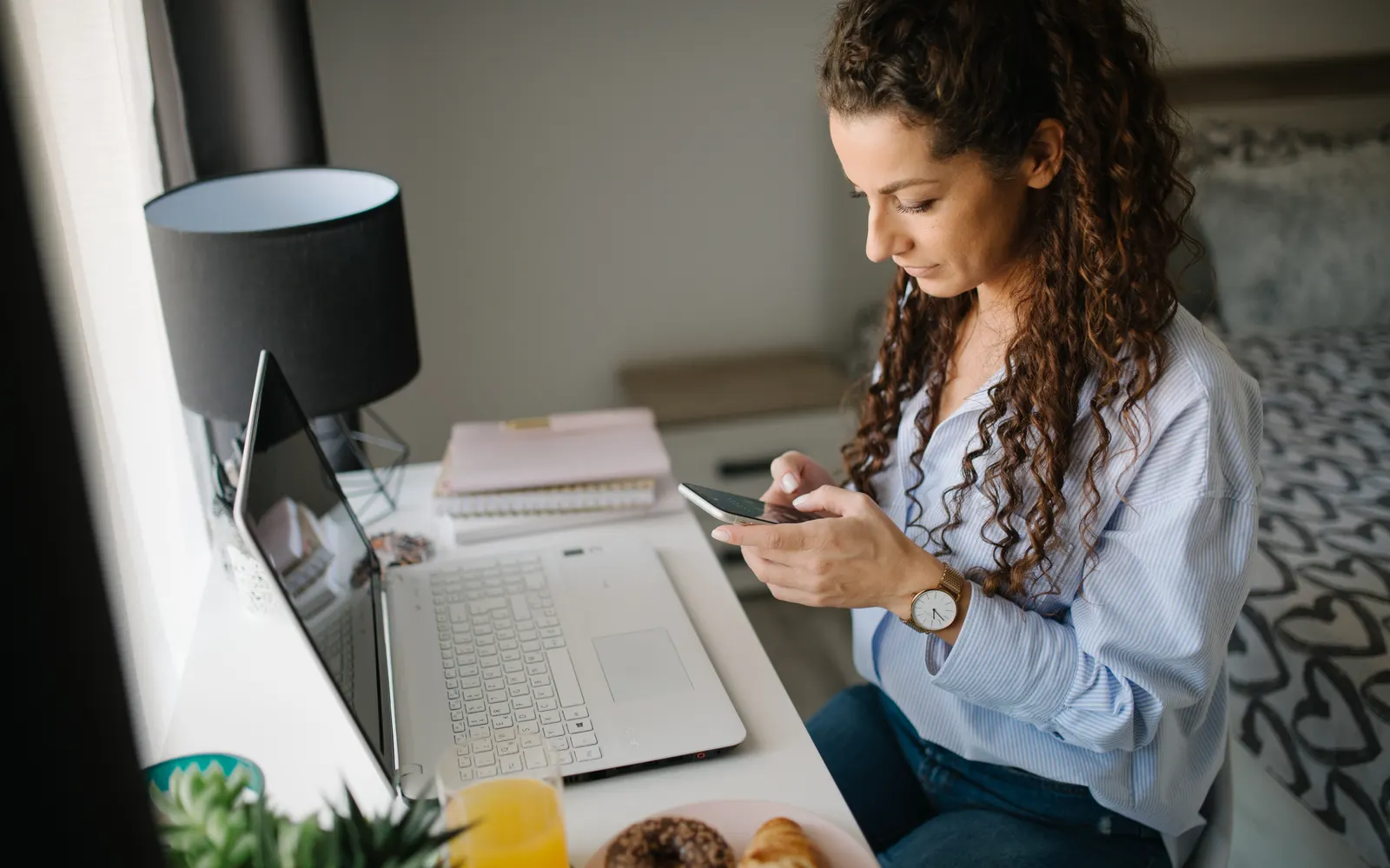 Woman checking her smartphone at a desk with a laptop and breakfast pastries
