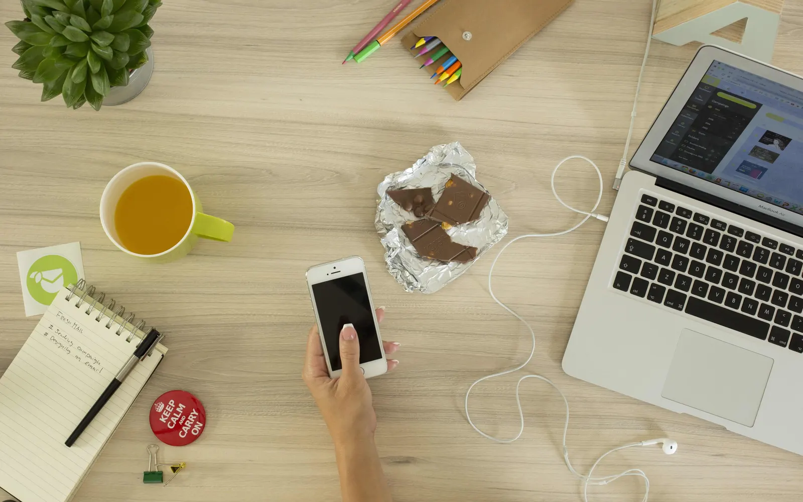 Top-down view of a hand holding a smartphone on a desk beside a laptop and pencils