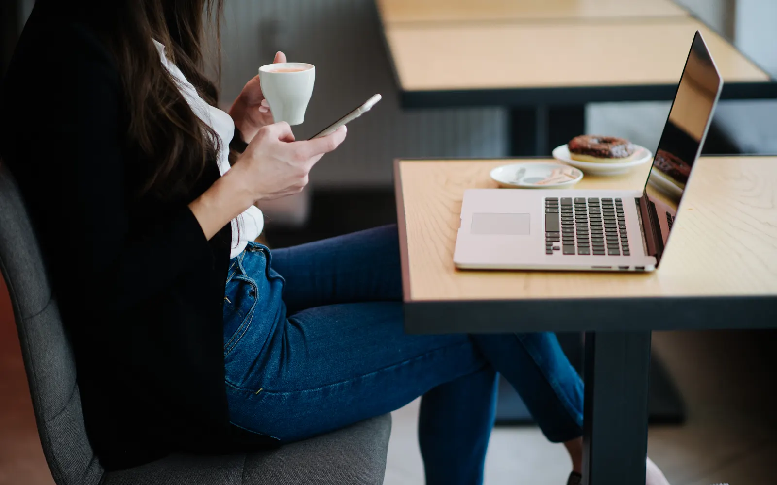 Woman in a café holding a coffee cup and a smartphone, laptop open on the table