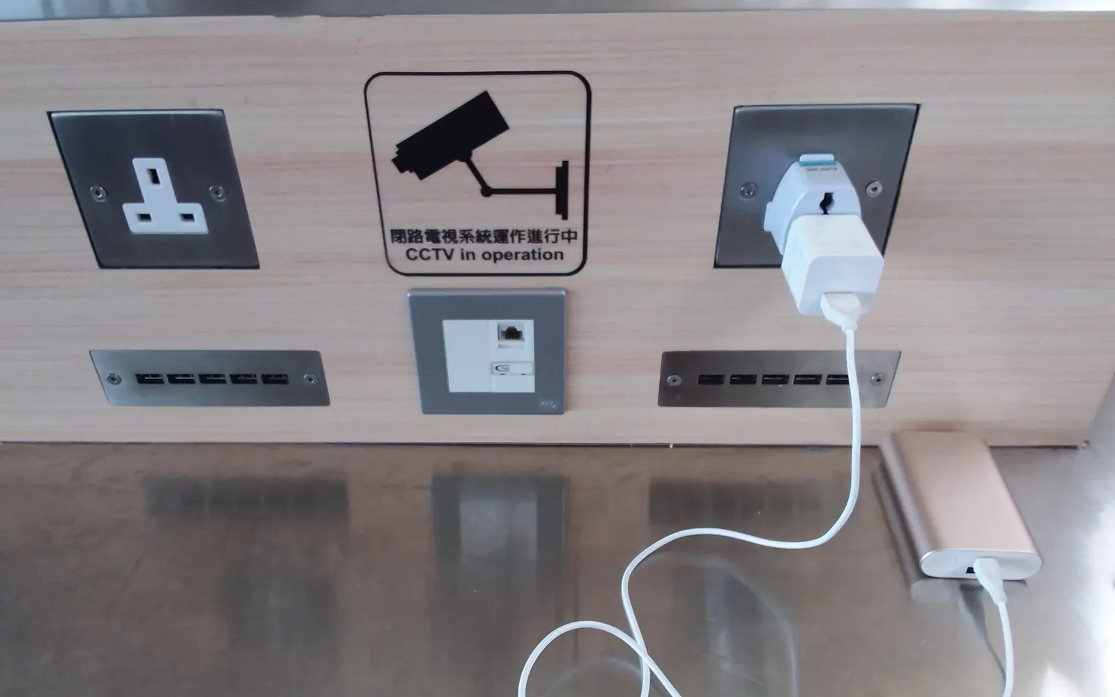 Phone charger plugged into a public charging point below a CCTV sign at an airport