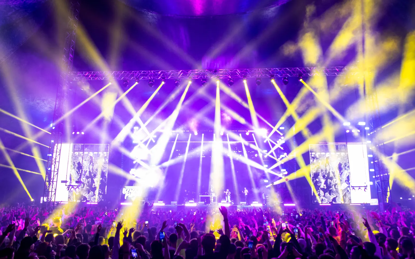 Crowd facing a festival stage lit with purple and yellow beams at night