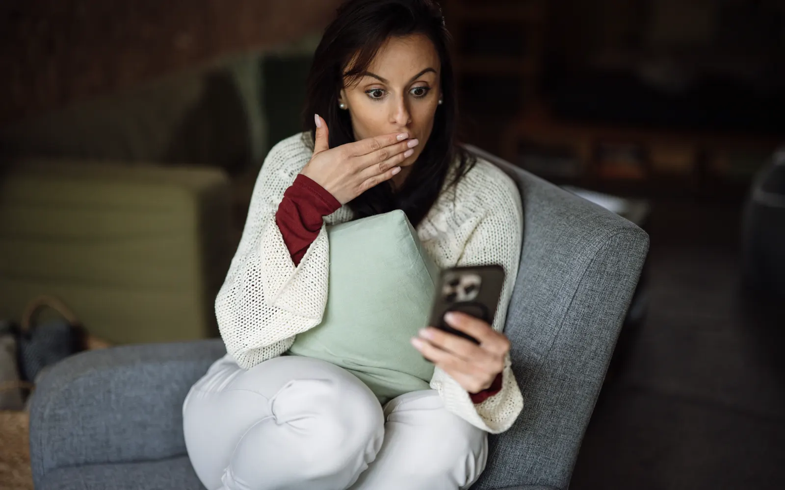 Woman on a sofa covering her mouth in surprise as she looks at a smartphone